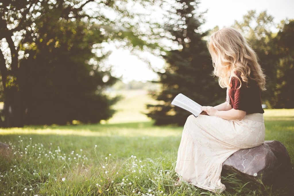 Blond lady reading a book, setting on a rock outside on green landscape for self-care