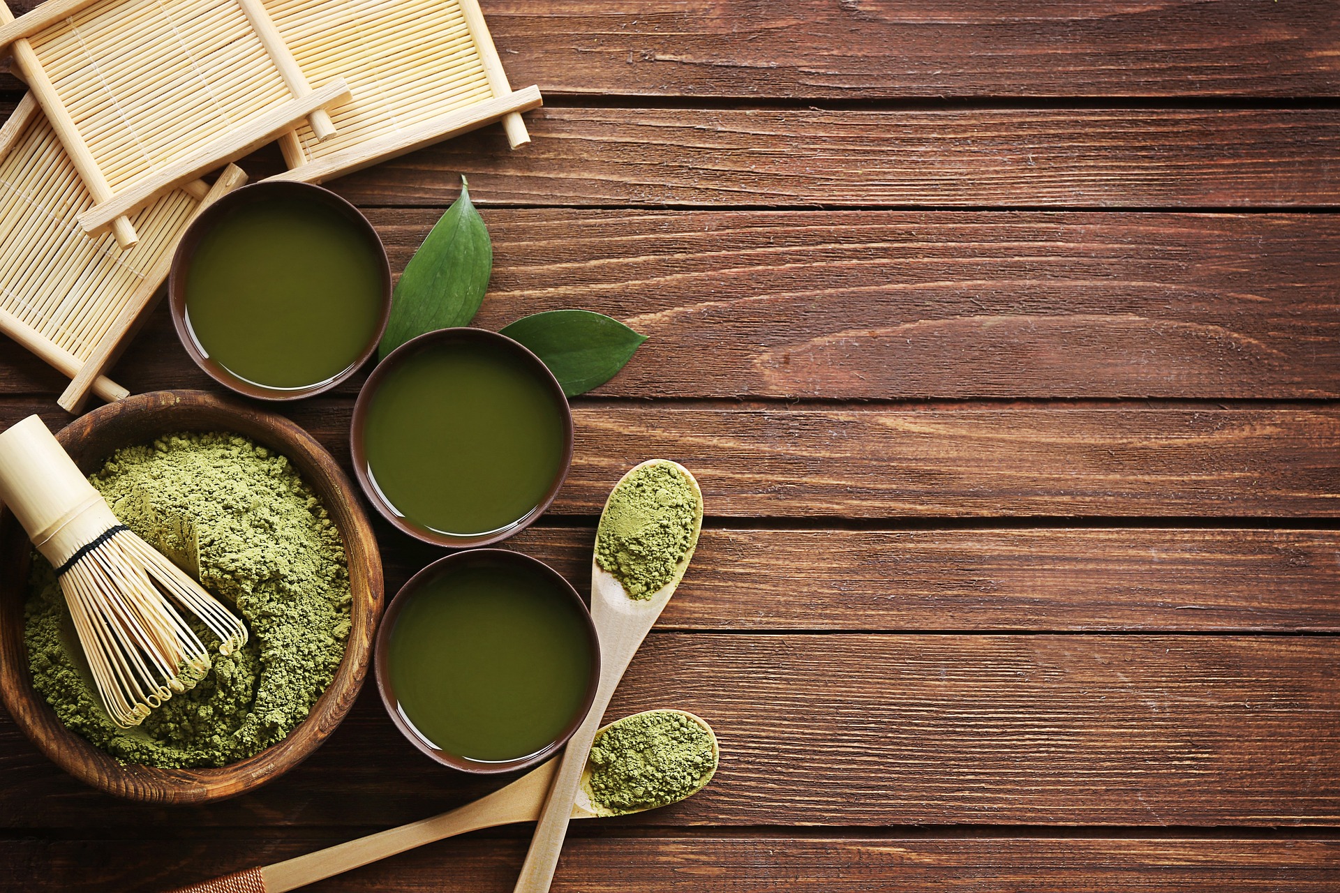 Morning routine with matcha powder, three cups of matcha drink and two wood spoons on a wood table