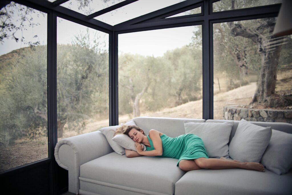Lady having a nap on a grey sofa inside a house with big windows and nature landscape