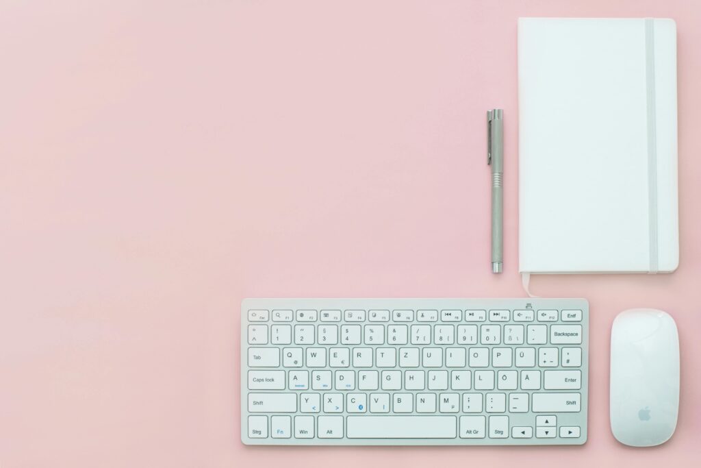 White keyboard, notebook. mouse and pen together with noitebook on a pinck background