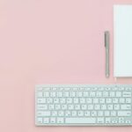 White keyboard, notebook. mouse and pen together with noitebook on a pinck background