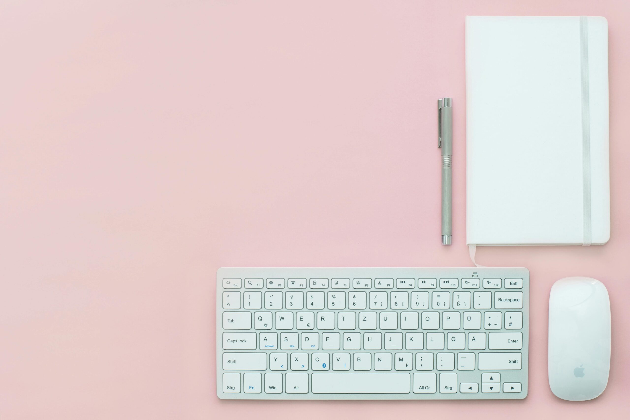 White keyboard, notebook. mouse and pen together with noitebook on a pinck background